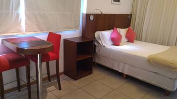 Una habitación de hotel pequeña con una cama individual, una mesita de noche, una silla y una ventana con cortinas.