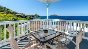 Un espacio al aire libre techado con mesa y sillas, con vista al mar, vegetación y una isla distante.