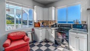 Una cocina con armarios blancos, piso a cuadros negro y blanco, una silla roja y vista al mar a través de amplias ventanas.