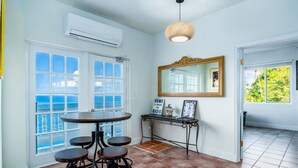 A room with a large window overlooking a beach, a round table with stools, a mirror, and a sign that reads "HOME IS WHERE THE BEACH IS".