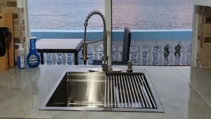 Une cuisine dotée d’une grande fenêtre donnant sur la mer, d’un évier en acier inoxydable et d’un tableau décoratif placé au-dessus de la fenêtre.