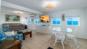 A spacious living room with a sofa, coffee table, dining table with chairs, a TV, and a view of the ocean through large windows.