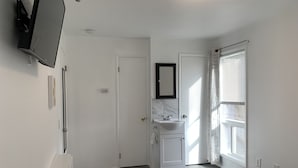 A bedroom with a bed, a bedside table, a TV mounted on the wall, a bathroom with a sink and mirror, and a window with blinds. - Central Bellevue Guesthouse  (Bellevue)