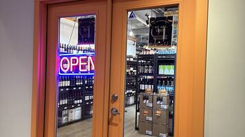 L’entrée d’une boutique de boissons alcoolisées, avec une enseigne néon indiquant « OUVERT », un panneau indiquant la présence d’un système de sécurité et une enseigne en bois au-dessus de la porte.