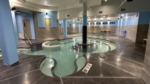Ein überdachter Schwimmteich mit kreisförmigem Design, umgeben von Wänden mit blauen und beigen Fliesen sowie einer Decke mit sichtbaren Leuchten.