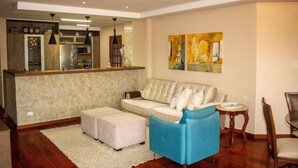 A modern living room with a sofa, ottoman, and a blue armchair. A kitchen area with a refrigerator and a dining table is visible in the background. - Apartamento Morro do Elefante (Campos do Jordão)