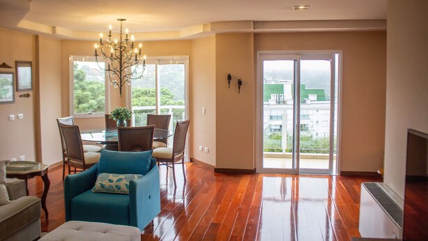 A spacious living room with a wooden floor, a dining area with a glass table, and a large sliding glass door leading to a balcony. - Apartamento Morro do Elefante (Campos do Jordão)