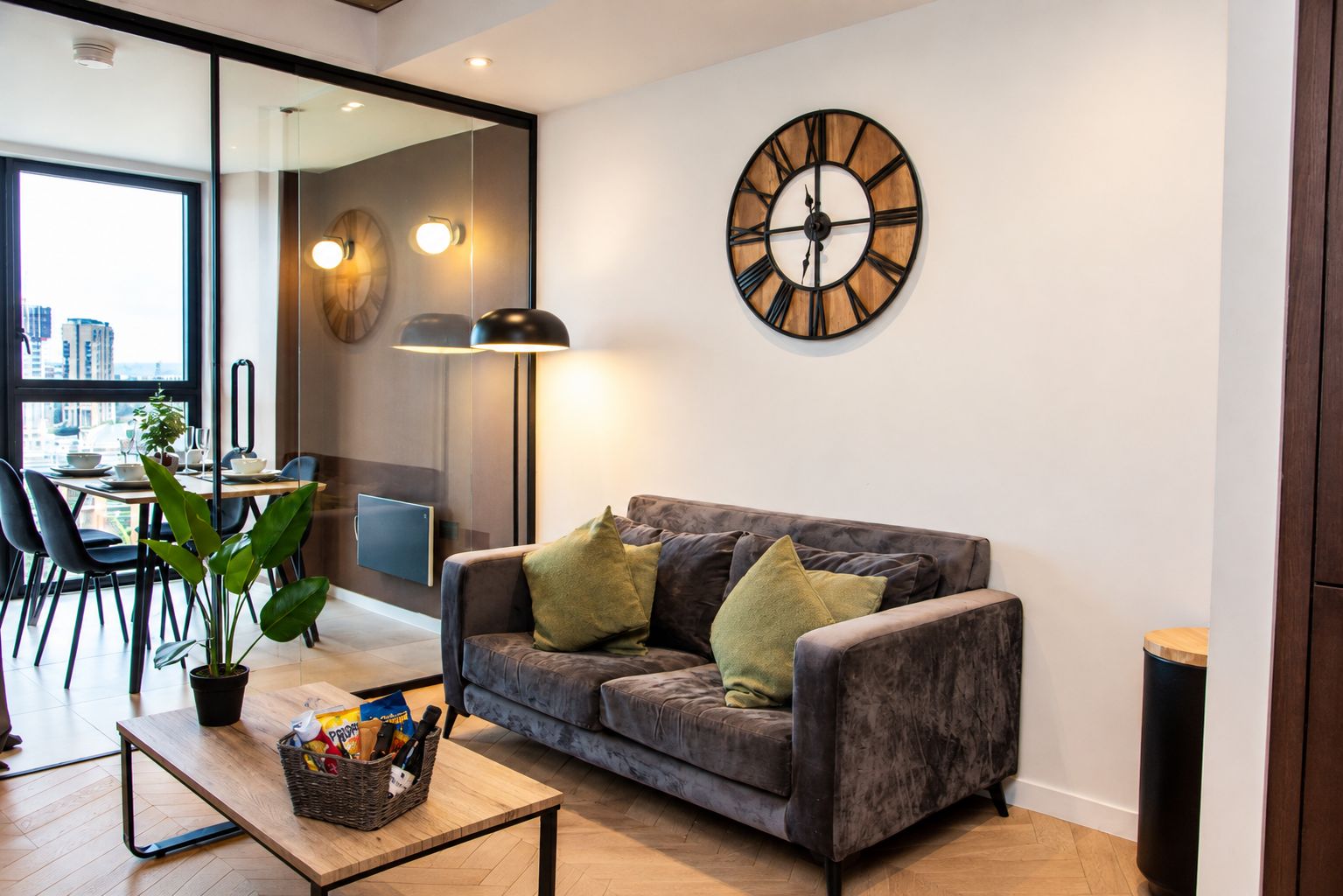 A modern living room with a grey sofa, a round wall clock, and a dining area with a glass partition.