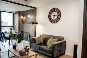Una sala de estar moderna con un sofá gris, un reloj de pared grande y una zona de comedor con una mesa y sillas.