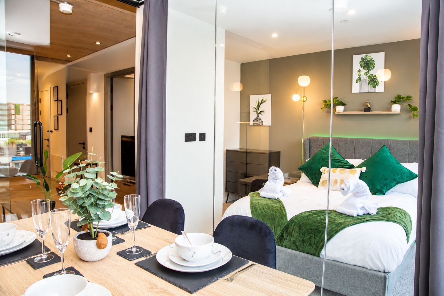 Une chambre d’hôtel moderne, équipée d’un lit, d’un coin repas avec une table dressée pour deux personnes, et offrant une vue à travers une paroi en verre.