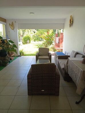 Una habitación con un sofá de mimbre, una mesita pequeña y una cesta de mimbre. Se ve un jardín a través de un ventanal.