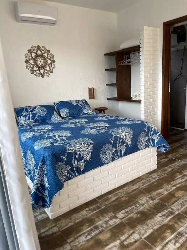 Camera da letto con testiera in mattoni bianchi, copripiumino floreale blu, climatizzatore a parete e mensola in legno con uno specchio.