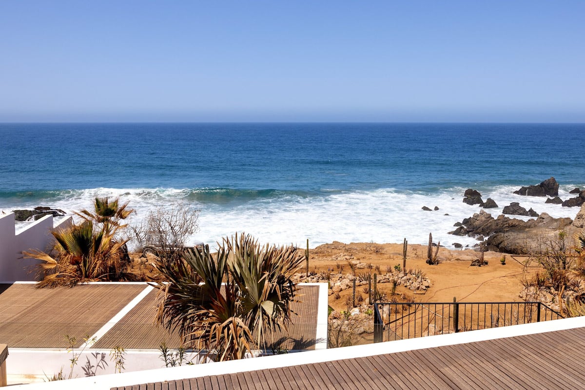 A coastal view with a sandy beach, rocky outcrops, and a wooden deck.
