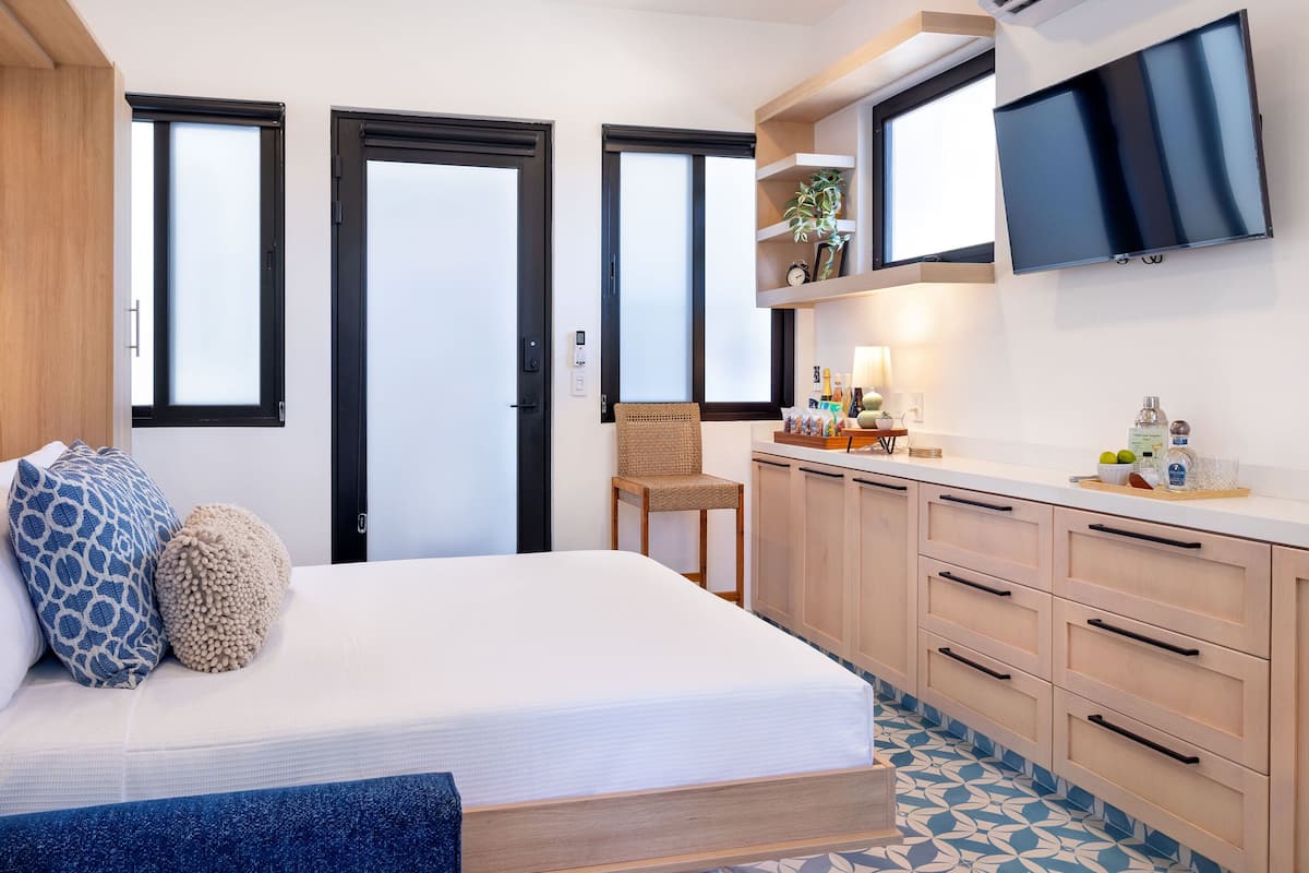 A modern bedroom with a bed, bedside tables, a chair, a TV, and a kitchen area with cabinets and a countertop.