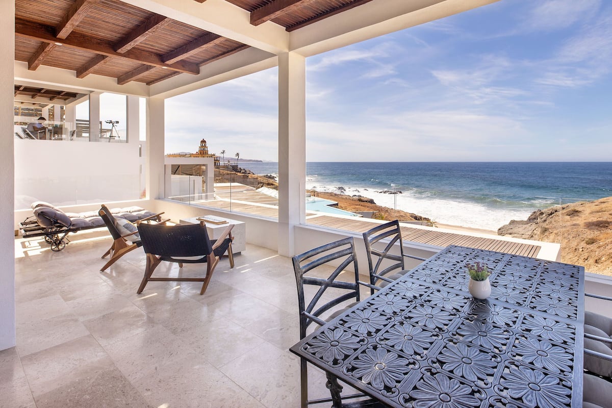 A terrace with a table and chairs overlooking a beach and ocean.