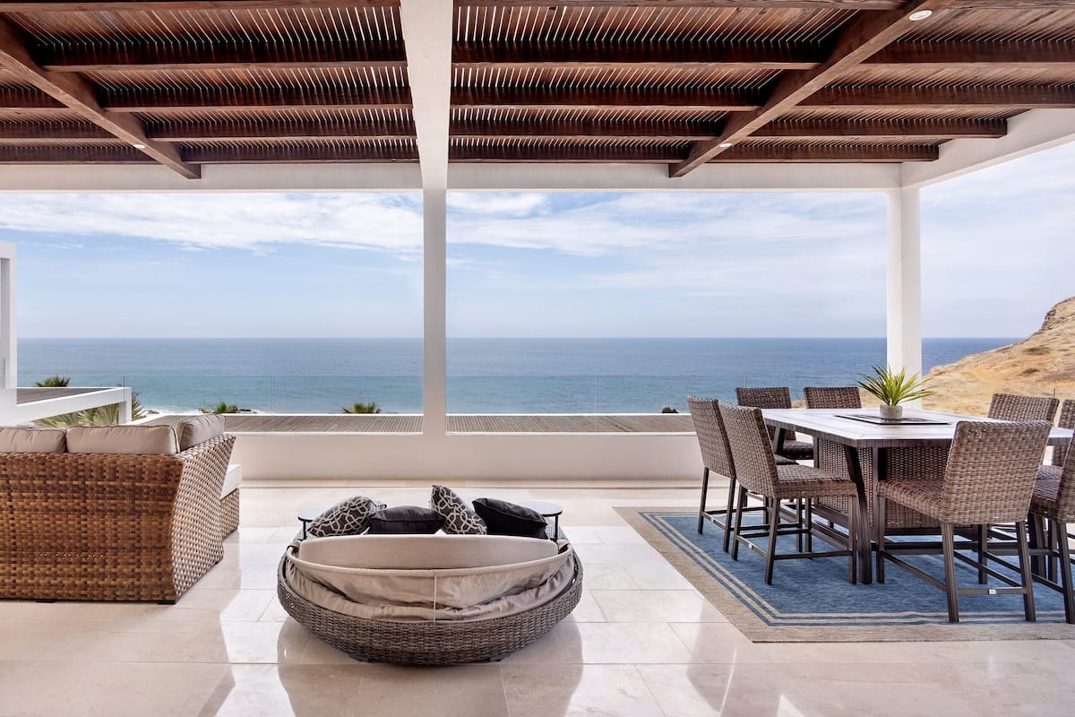 A covered outdoor seating area with a view of the ocean, featuring wicker furniture and a dining table.