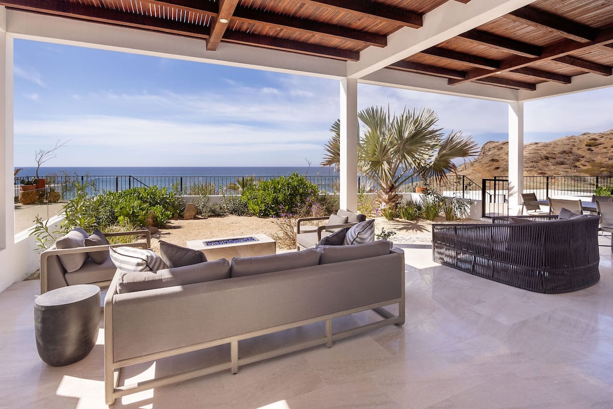 A covered outdoor seating area with a view of the sea, furnished with sofas and chairs, and a dining table.