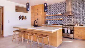 A modern kitchen with a large island, wooden stools, a stainless steel stove, and patterned backsplash.