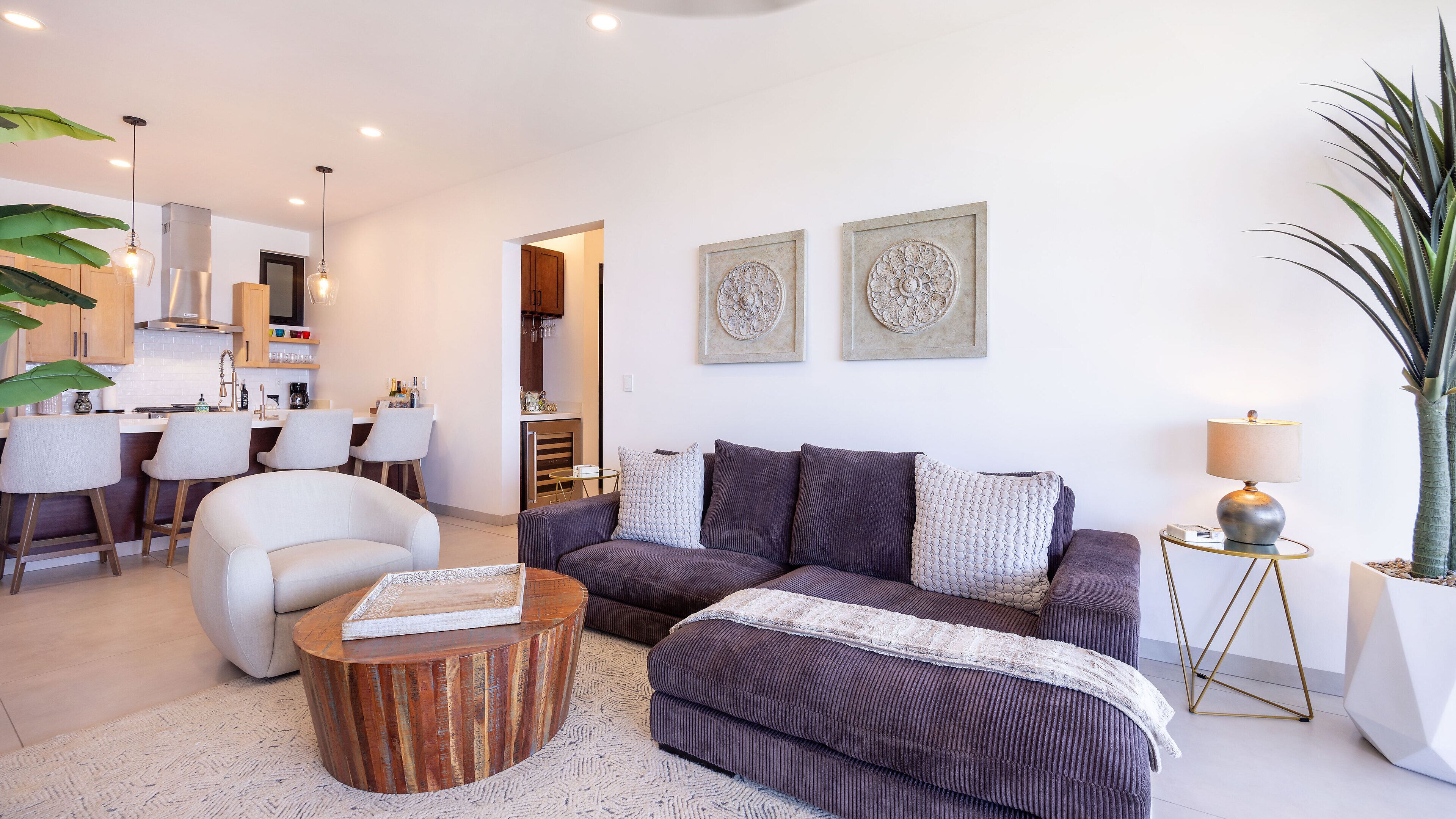 Un salon moderne avec un canapé, une table basse et des coussins décoratifs. On aperçoit un coin repas avec des chaises et, au fond, une cuisine.