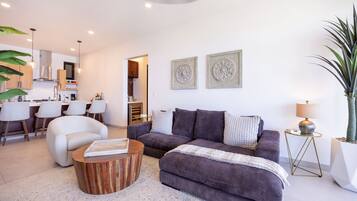 Un salon moderne avec un canapé, une table basse et des coussins décoratifs. On aperçoit un coin repas avec des chaises et, au fond, une cuisine.