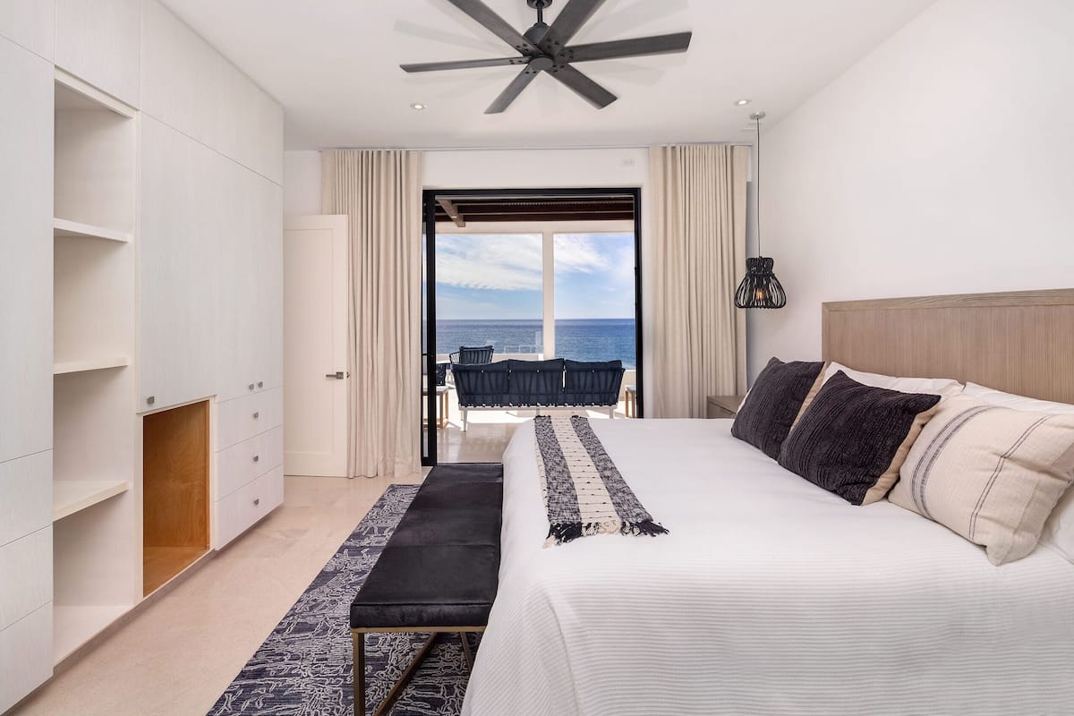 A bedroom with a large bed, a ceiling fan, a view of the ocean, and a balcony with a chair.