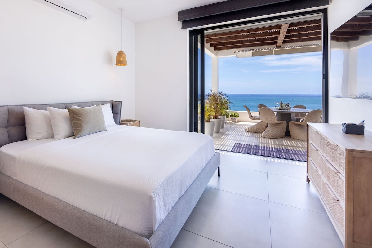 A bedroom with a large bed, a balcony with a view of the sea, and a wooden desk.
