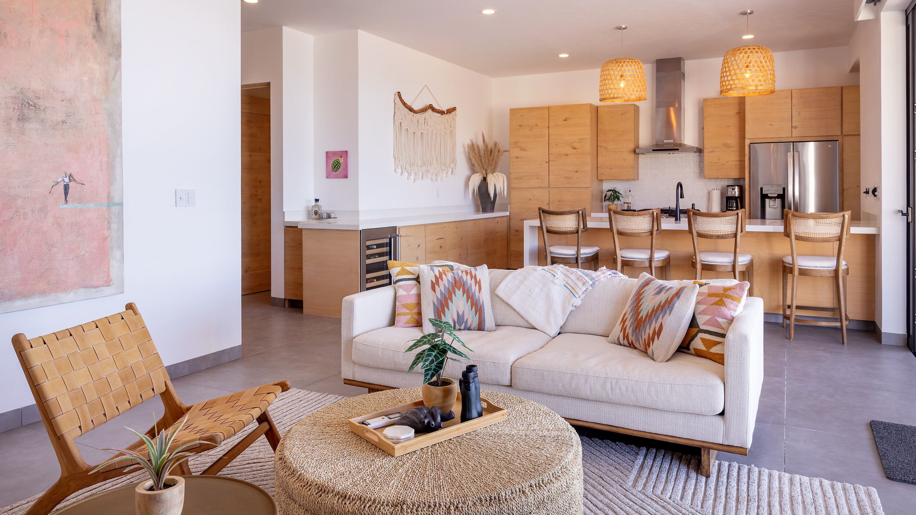 Ein modernes Wohnzimmer mit Sofa, Sesseln und Couchtisch. Es gibt eine Küchenzeile mit Holzschränken und einem Kühlschrank.