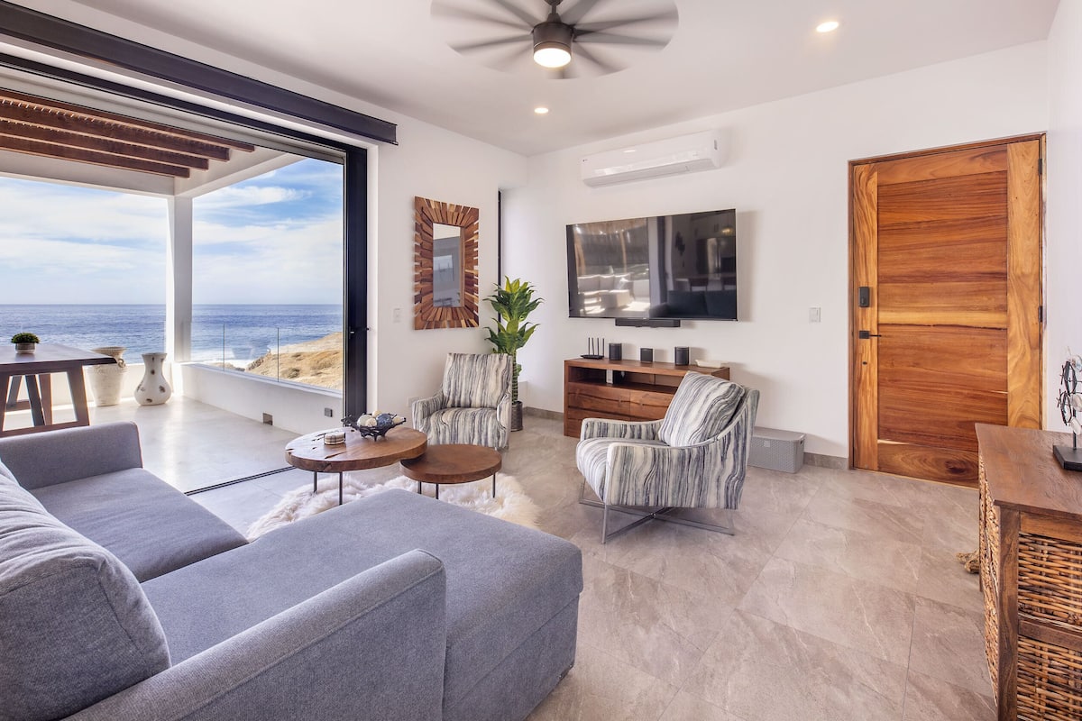 A modern living room with a large sofa, a coffee table, and a TV. There is a wooden door and a large window overlooking the ocean.