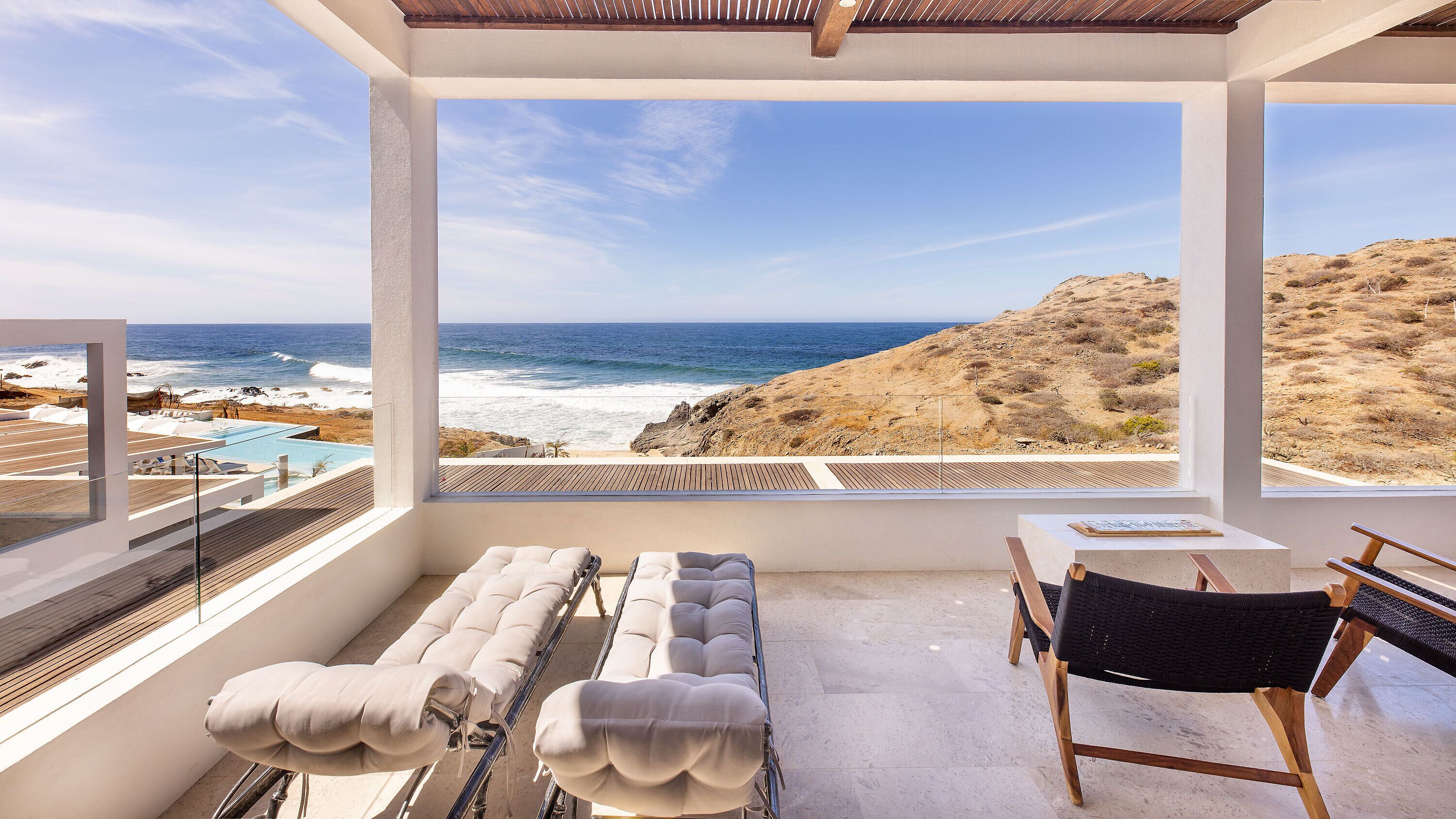 Un balcón con vista al océano, una piscina y una zona de estar con cojines y una mesa.