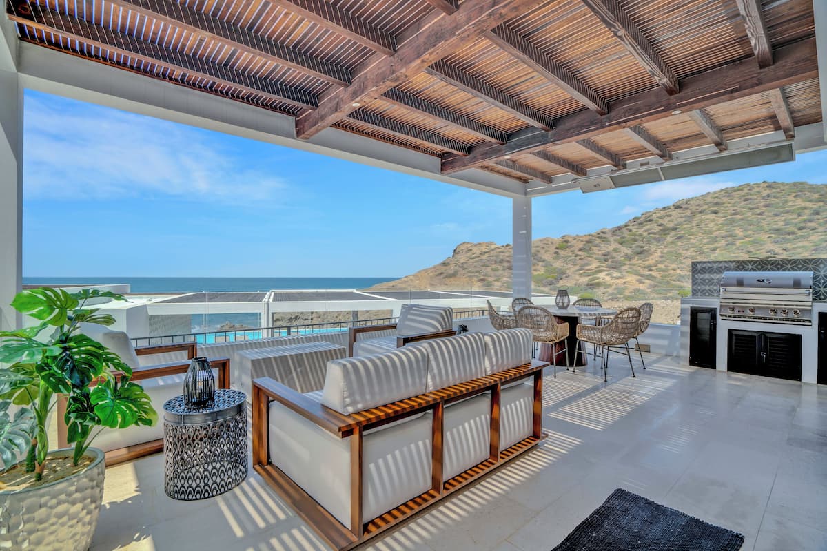 A modern outdoor patio with a wooden ceiling, a seating area with cushions, a grill, and a view of the ocean.