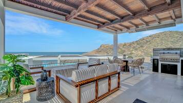 A modern outdoor patio with a wooden ceiling, a seating area with cushions, a grill, and a view of the ocean.