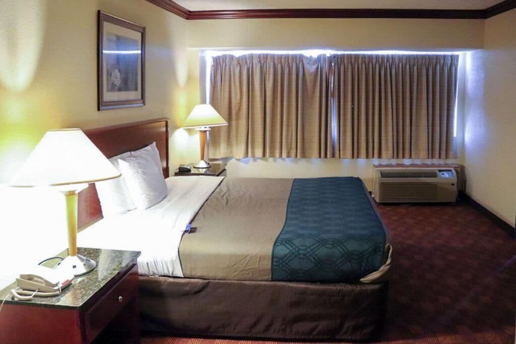 A hotel room with a bed, two bedside lamps, a window with curtains, an air conditioning unit, and a framed picture on the wall.