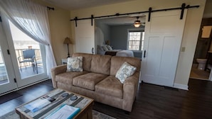 A living room with a brown sofa, a coffee table with magazines, a sliding door, and a view of a balcony. - Vineyard Villa overlooking 40 acres of Vineyards in Ohio Wine Country (Geneva)