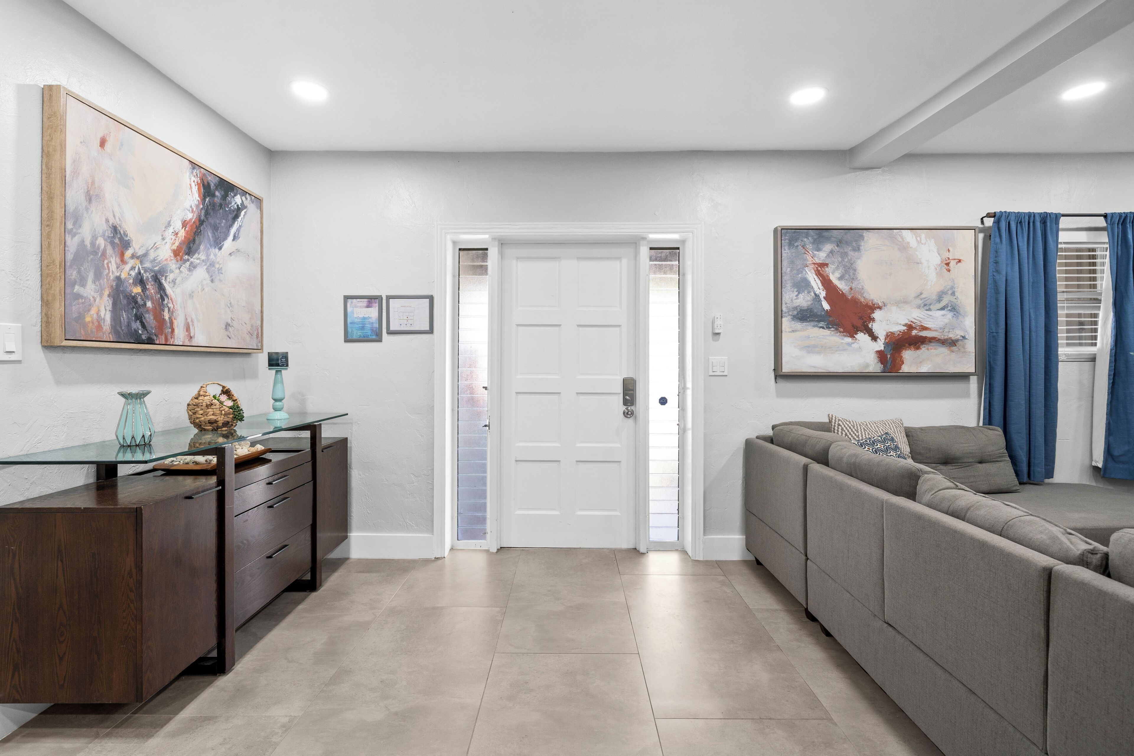 A modern living room with a glass coffee table, a dark wooden console, a grey sectional sofa, and abstract paintings on the walls.