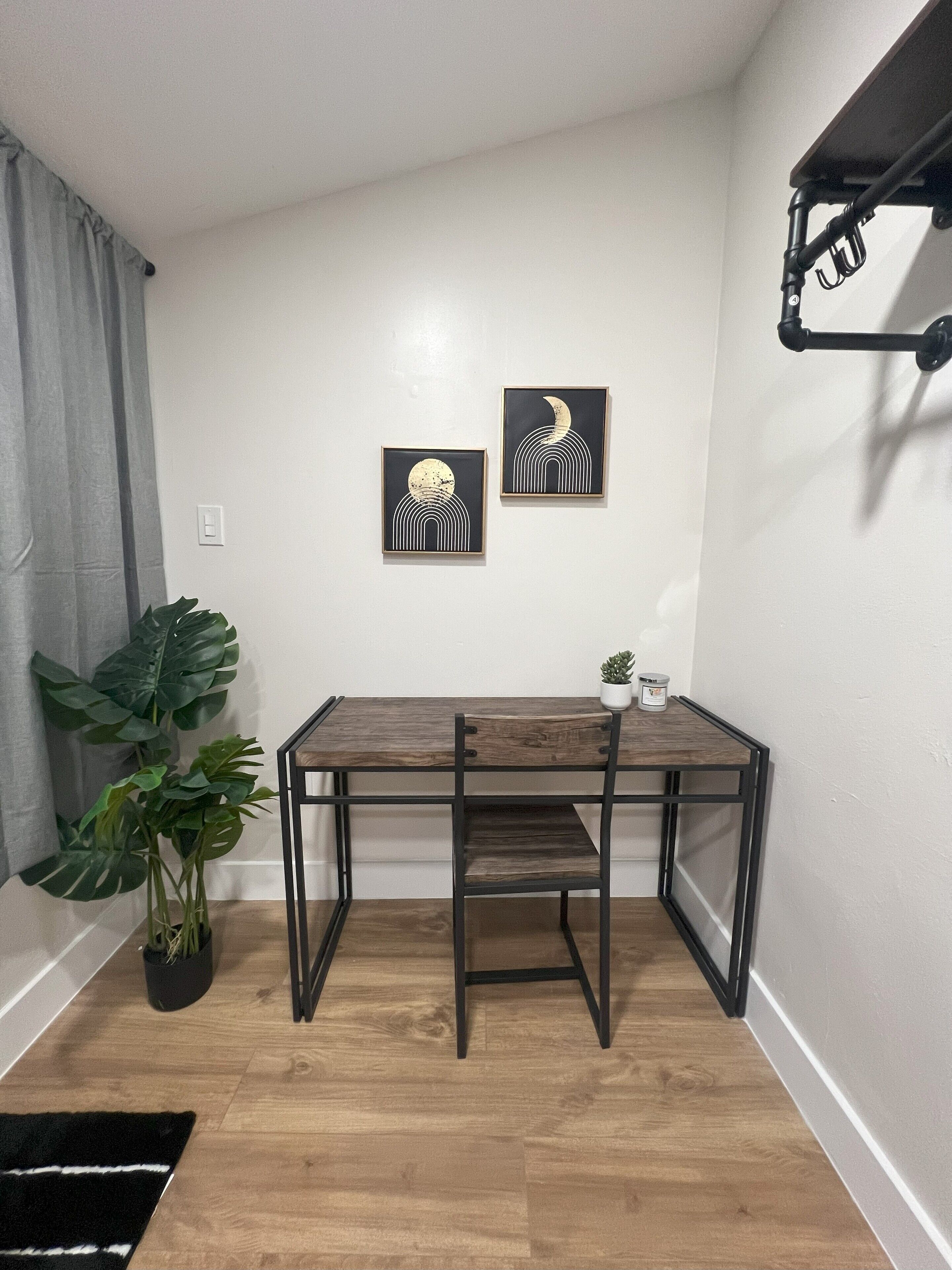 A compact room with a wooden table, two framed artworks, a potted plant, and a ceiling-mounted rack.