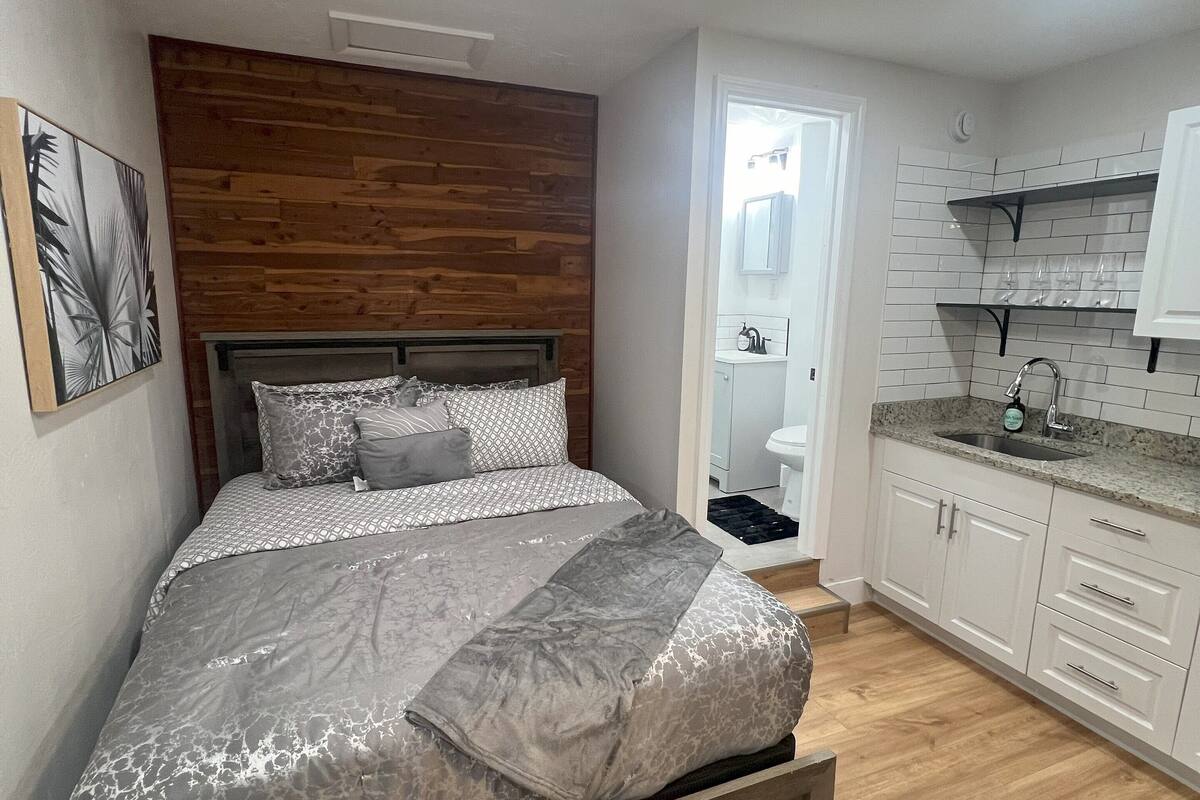 Ein Schlafzimmer mit einem hölzernen Kopfteil, einem Bett mit gemusterten Betttextilien und einem Nachttisch. Angrenzend an das Schlafzimmer befindet sich eine Küche mit weißen Schränken und einer Arbeitsplatte.