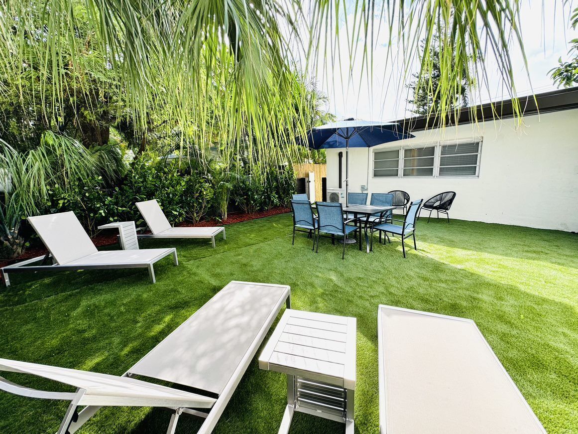 A well-maintained outdoor patio with white lounge chairs and a table, surrounded by lush greenery and a modern building in the background.