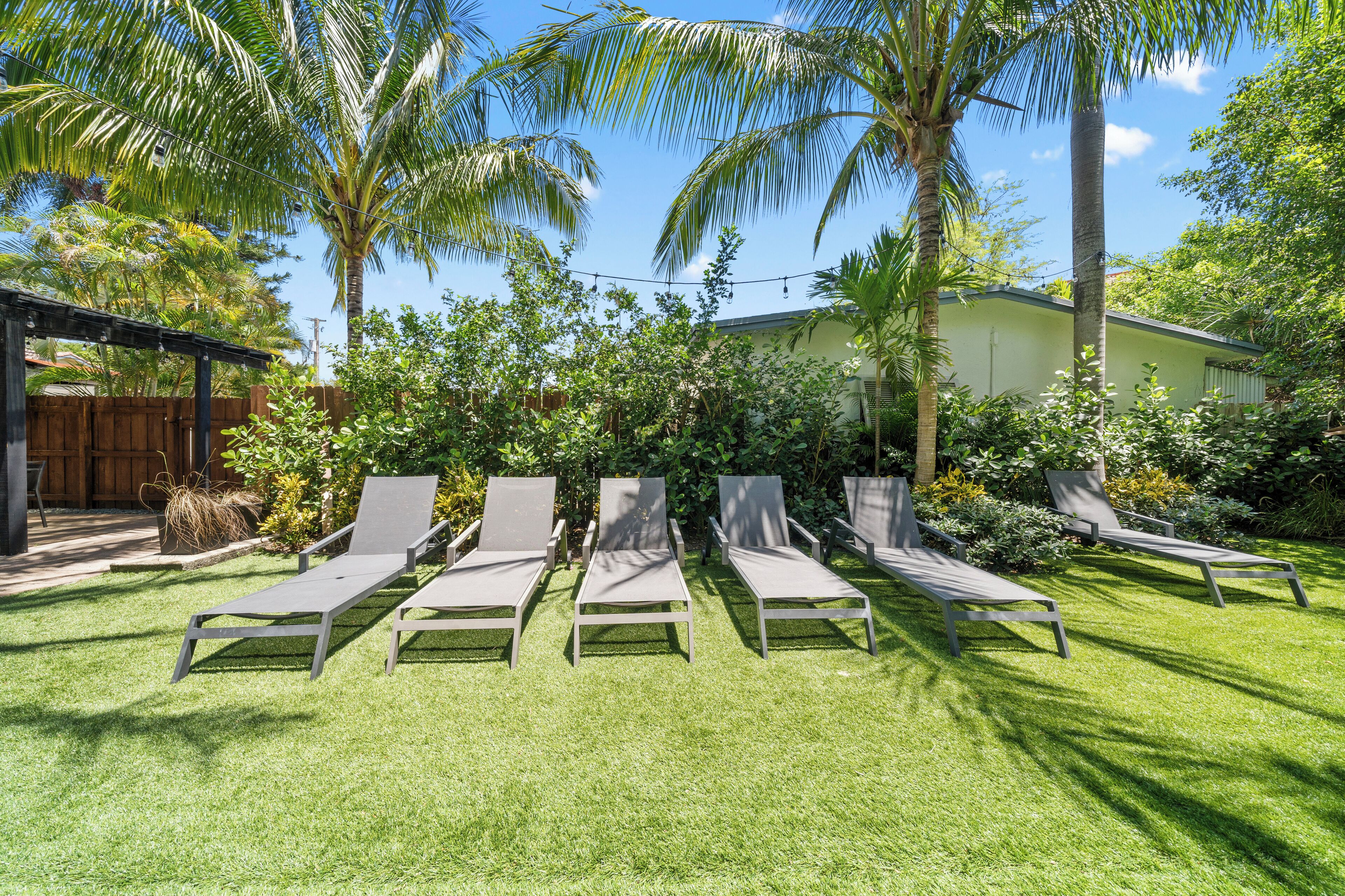 Un jardin avec cinq chaises longues disposées en rangée, entouré d’une végétation luxuriante et de palmiers.