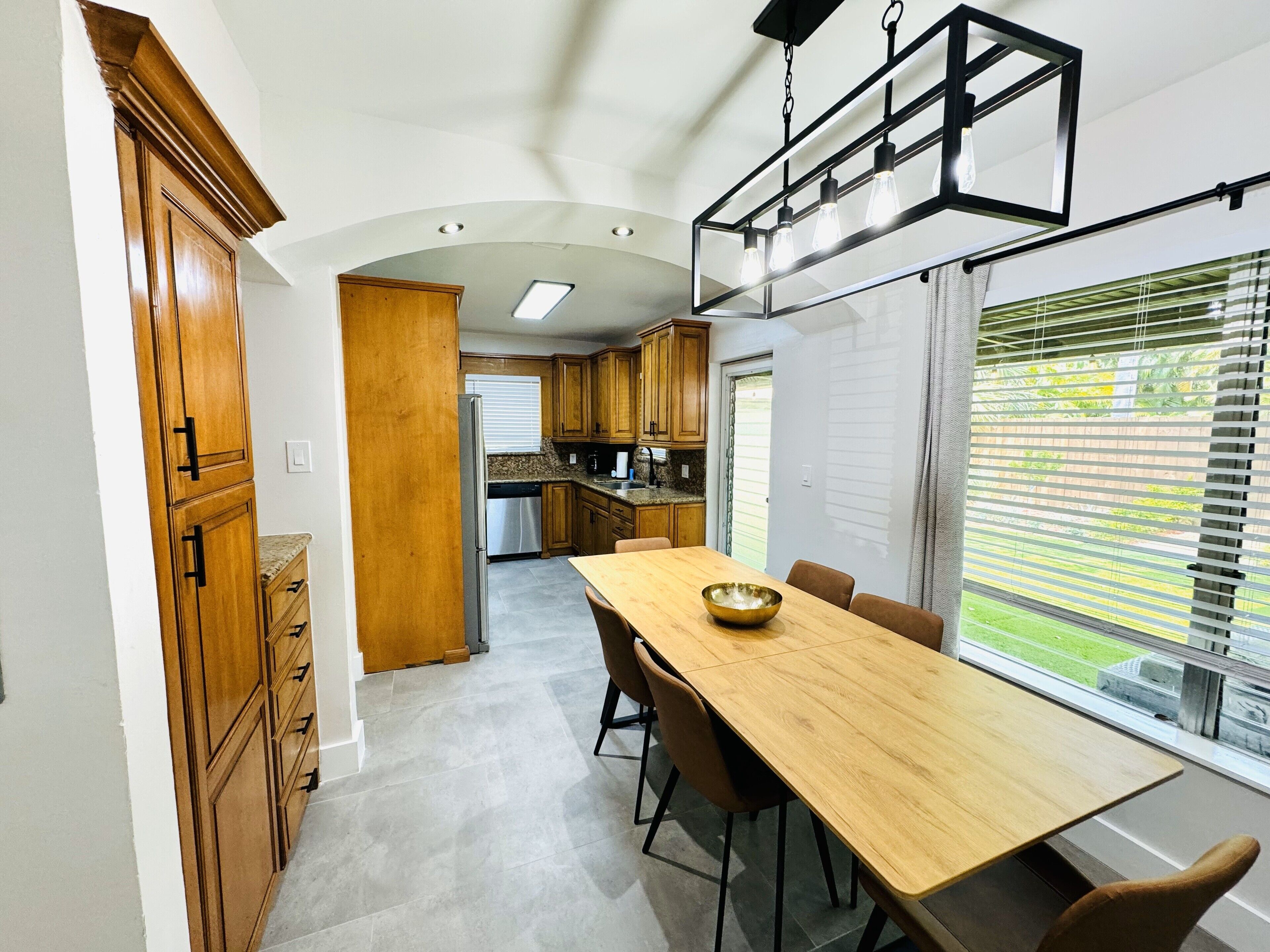 A modern kitchen with wooden cabinets, a stainless steel refrigerator, and a dining area with a long wooden table and chairs.