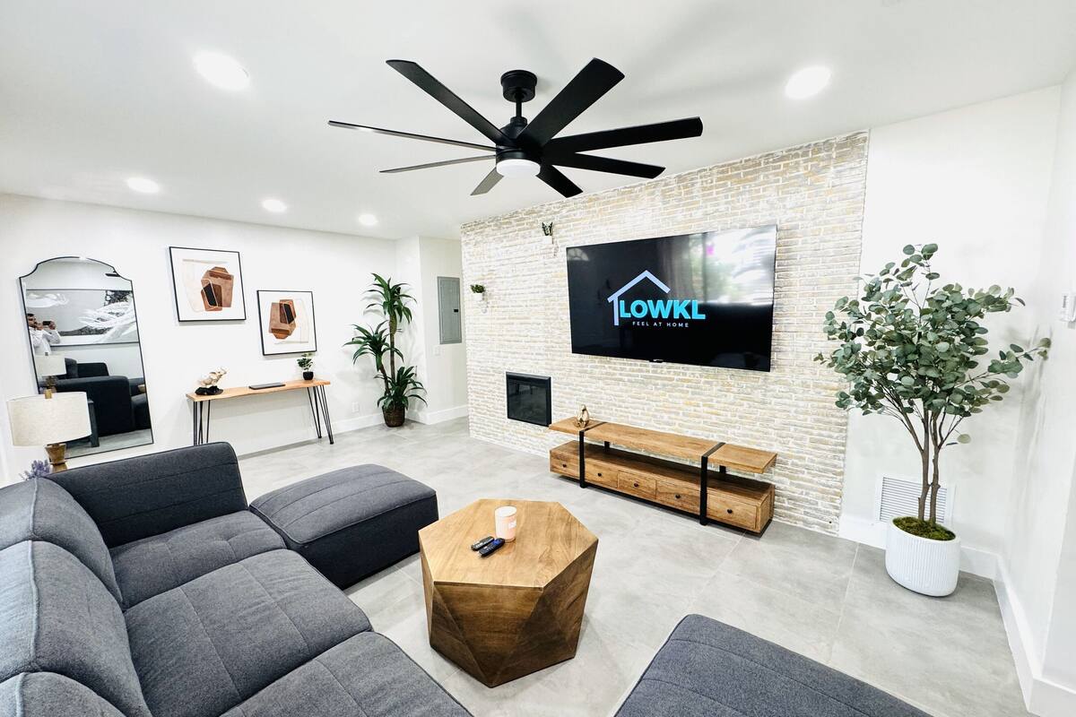 Ein modernes Wohnzimmer mit einem Ecksofa, einem Couchtisch, einem an einer Ziegelwand montierten Fernseher, einem Deckenventilator und einer Topfpflanze.
