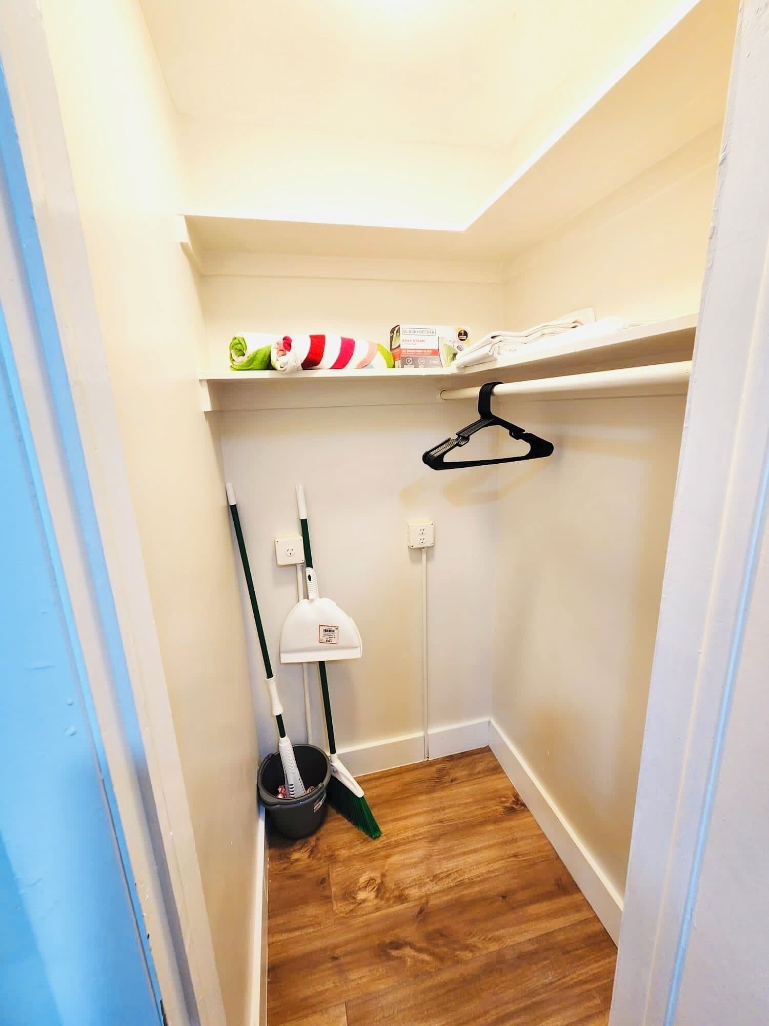 A narrow hallway with wooden flooring, a broom, a mop, and a hanging rack.