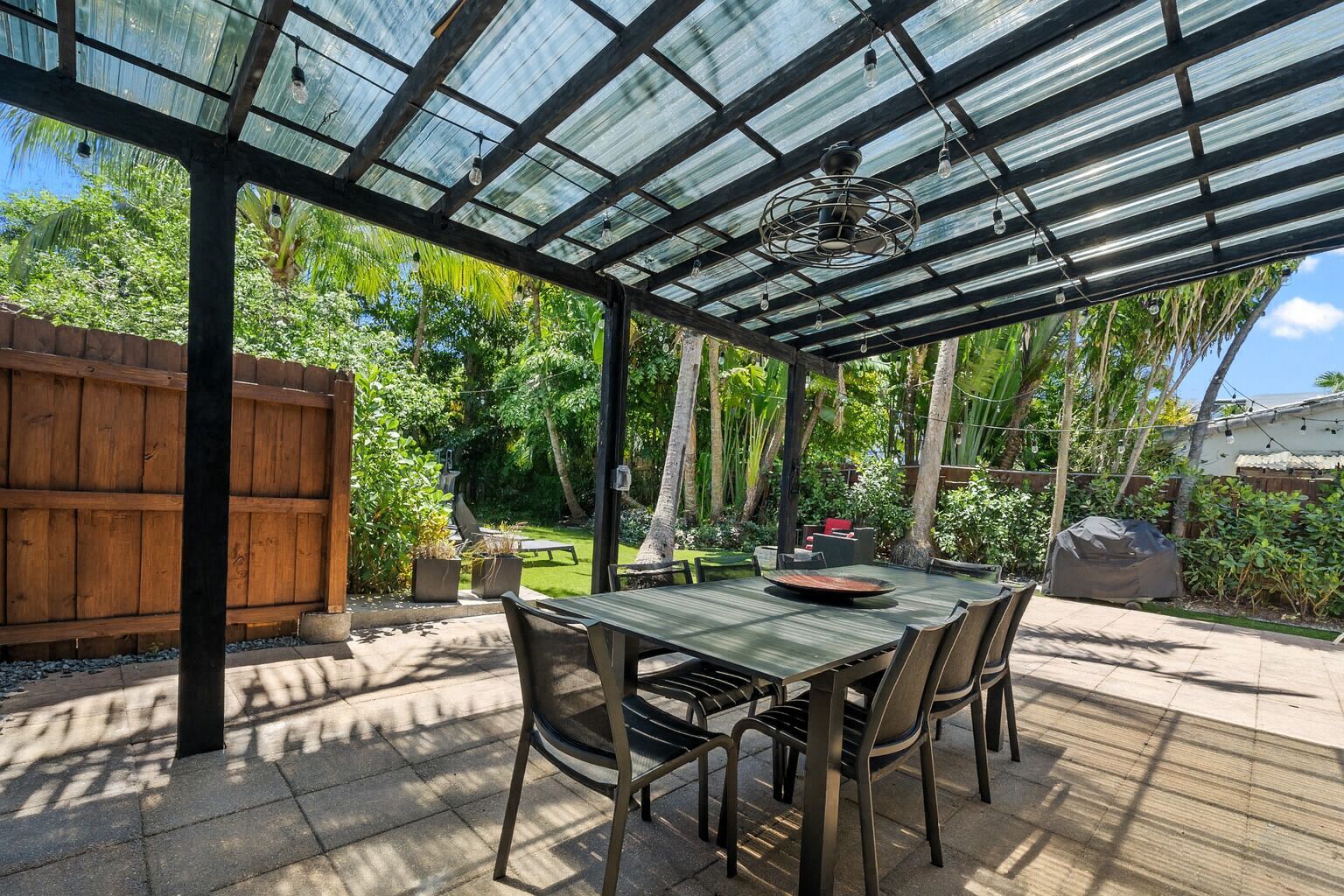 A covered patio with a wooden table and chairs, surrounded by a wooden fence and lush greenery.