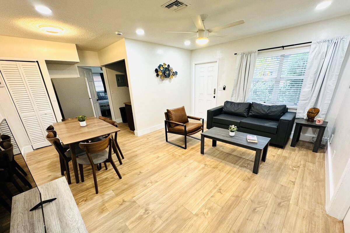 Ein modernes Wohnzimmer mit einem Holzesstisch, einer schwarzen Couch und einem Couchtisch. Es gibt einen Deckenventilator, ein Fenster mit Vorhängen und eine Tür zu einem anderen Raum.