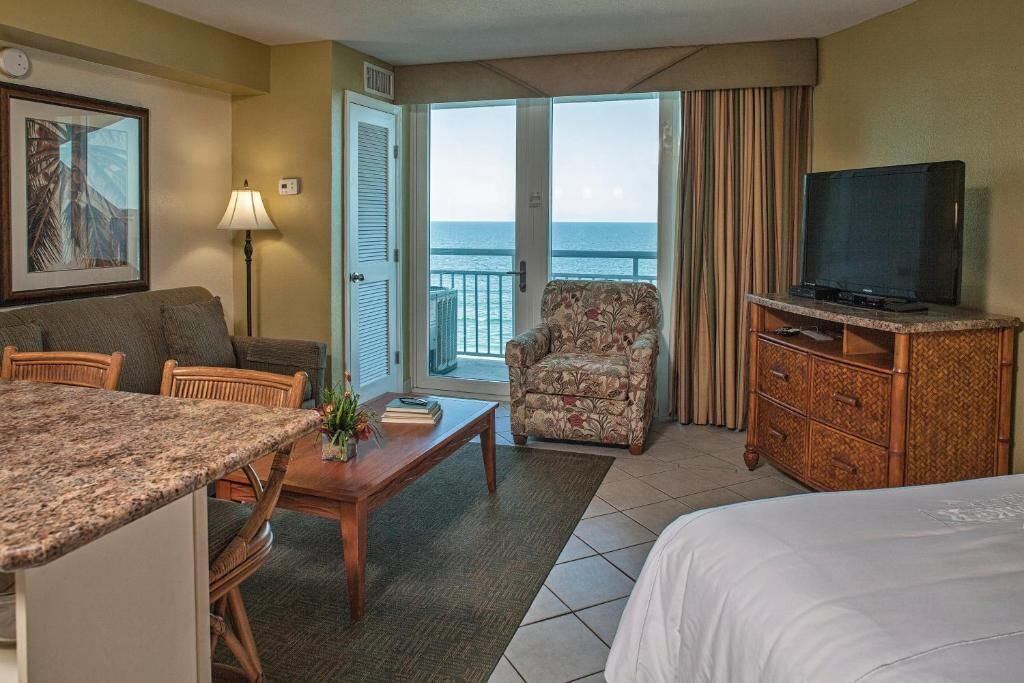 A hotel room with a bed, a television, a dining area with chairs and a table, a chair with a floral pattern, and a view of the ocean through the balcony doors.