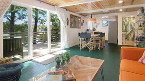 A spacious living area with a wooden floor, a dining table with chairs, a sofa, and a large window offering a view of trees.