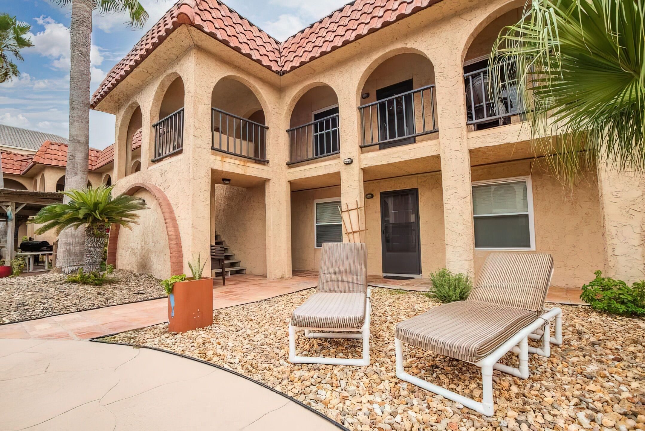 A building with a stucco exterior, red-tiled roof, and multiple balconies. There are two lounge chairs in the foreground.