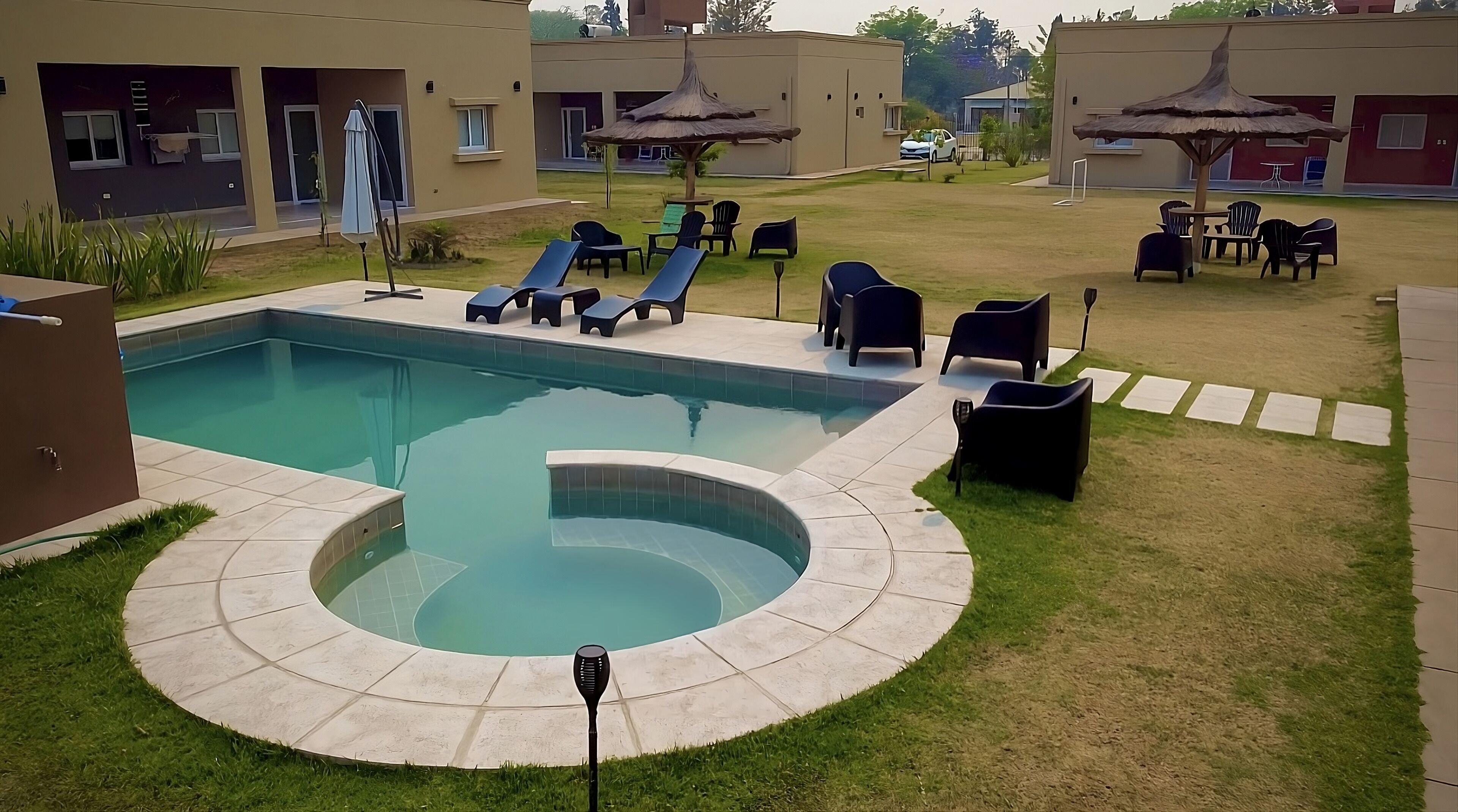 Une piscine dotée de chaises longues et de parasols, entourée d’une pelouse bien entretenue et d’un bâtiment comprenant plusieurs pièces.