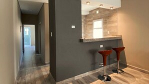 A modern hallway with a dark wall, a wooden floor, and a kitchen area with a countertop and two stools. - Entre Cerros Apart (Salta)