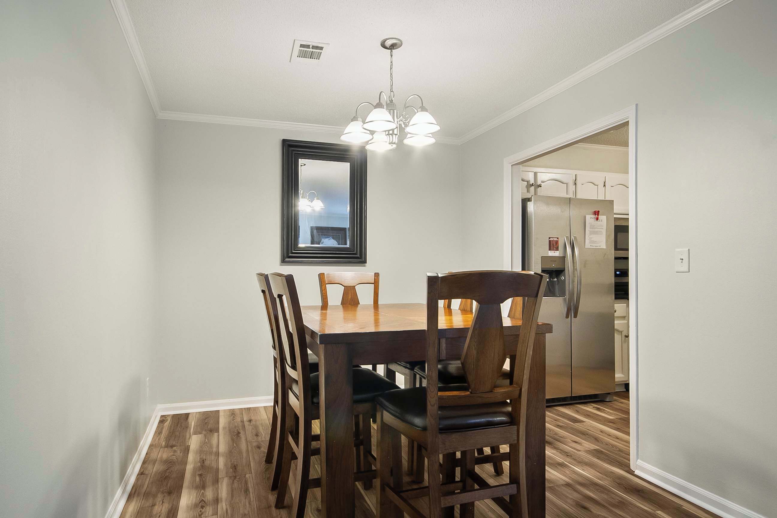 A dining area with a wooden table and chairs, a chandelier, and a view of the kitchen through an open door.