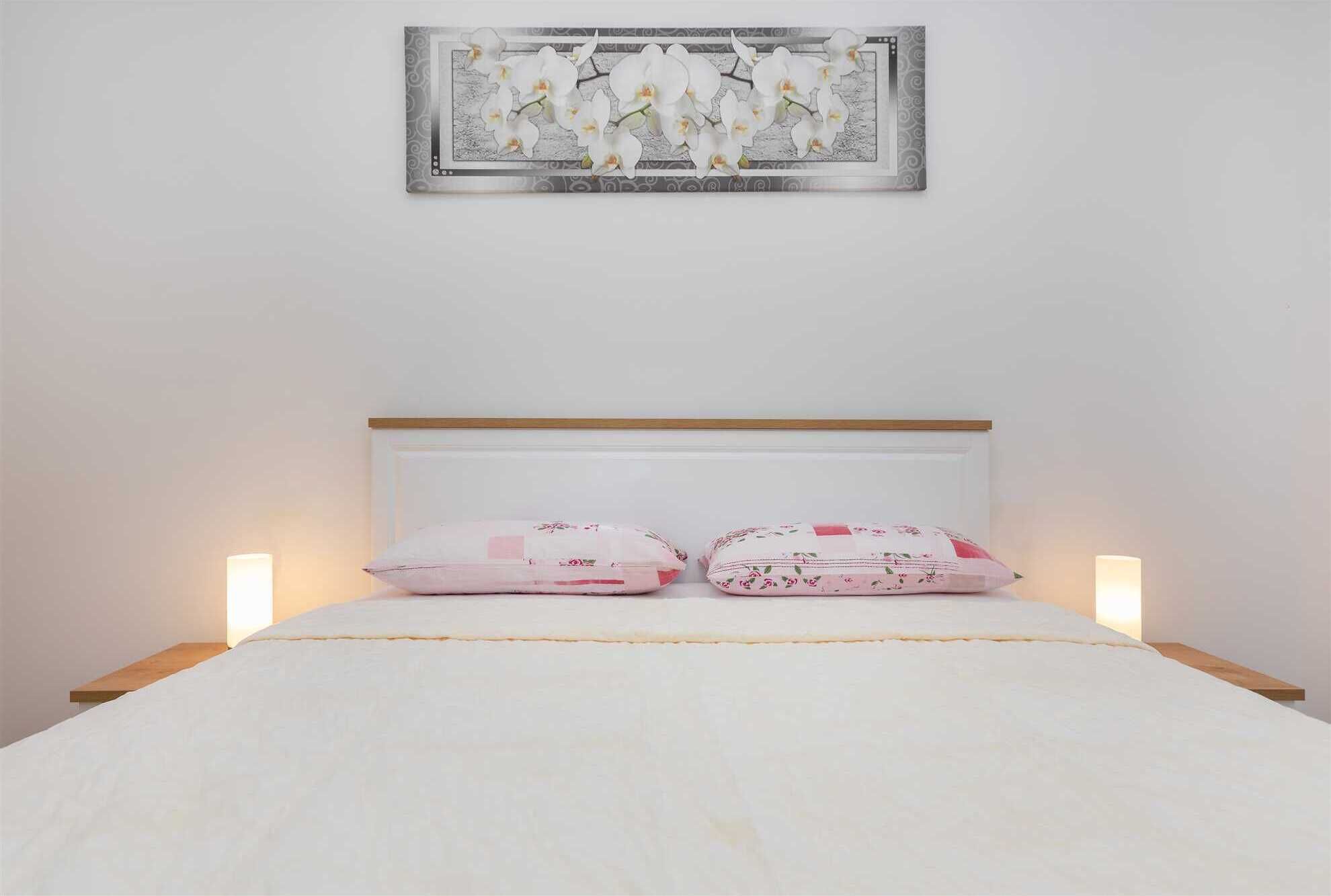 A bed with a white mattress and two floral-patterned pillows, a wooden headboard, and a framed picture of white orchids on the wall.